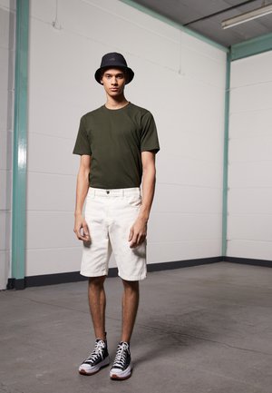 T-shirt vert olive à manches courtes, shorts blancs, chapeau seau noir et baskets montantes noires avec des accents blancs, se tenant dans un espace minimaliste.