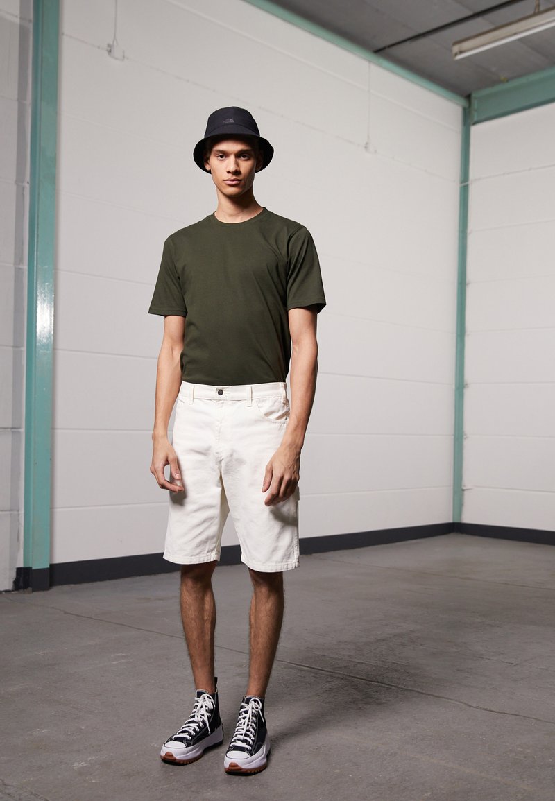 T-shirt vert olive à manches courtes, shorts blancs, chapeau seau noir et baskets montantes noires avec des accents blancs, se tenant dans un espace minimaliste.
