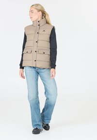 Beige vadderad väst med hög krage, framknappar och två ytters fickor, bar över en svart långärmad skjorta och blå jeans.