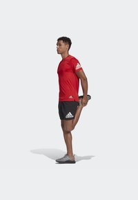 Maglietta sportiva rossa e pantaloni corti neri con logo Adidas bianco; la persona allunga una gamba all'indietro mentre indossa scarpe sportive grigie.