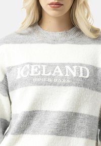 Un pull rayé gris et blanc avec un col rond, comportant "ICELAND" brodé en blanc, confectionné dans un mélange de laine doux et texturé.