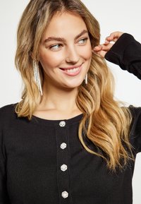 Fekete kötött pulóver kerek nyakkivágással, öt díszítő ezüstgombbal és hosszú ujjakon. A modellen hullámos haj és hosszú fülbevalók találhatók.