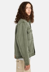 Chaqueta de denim verde con corte relajado, que presenta un cierre de botones, dos bolsillos en el pecho y un acabado texturizado. Llevada sobre una camiseta gris claro.