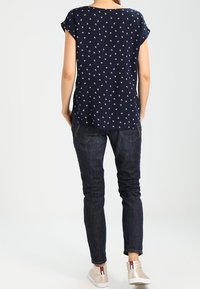 Marinblus med kort ärm i vitt ankarmönster, tillsammans med mörka jeans och ljusa sneakers. Bakvy visar en avslappnad passform och design.