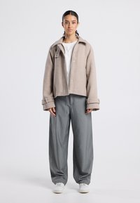 Young woman with dark hair in beige jacket, white shirt, loose gray pants, and white sneakers standing against a plain light background.