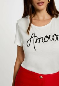 Witte korte mouwen t-shirt met zwart geborduurd "Amour" tekst. Gekleed met hoge taille rode broek. Glad materiaal met een ronde halslijn.