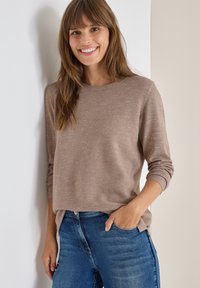 Hellbrauner, strukturierten Pullover mit runder Ausschnitt und langen Ärmeln, kombiniert mit dunkelblauen Jeans, vor einem neutralen Hintergrund.
