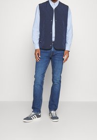 Marineblau gesteppte Weste mit Knopfleiste, getragen über einem hellblauen Hemd. Ein Paar blaue Jeans und graue Sneakers mit weißen Streifen.