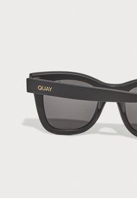 Lunettes de soleil noires avec une finition mate, dotées d'une monture légèrement courbée, de verres foncés et d'un logo "QUAY" doré sur la branche.