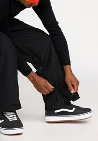 Pantalons noirs imperméables avec des chevilles zippées, associés à des baskets noires avec des accents blancs, sur un fond clair.