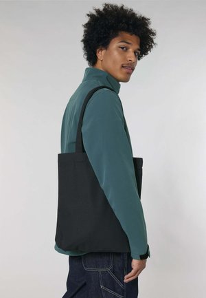 Zwarte tote bag met lange handvatten, gemaakt van stevig stof, hangt over de schouder van een persoon die een groene jas draagt tegen een lichte achtergrond.