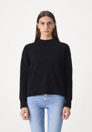 Black fuzzy pullover sweater with a high neckline and long sleeves, paired with light blue jeans featuring a button-up front.