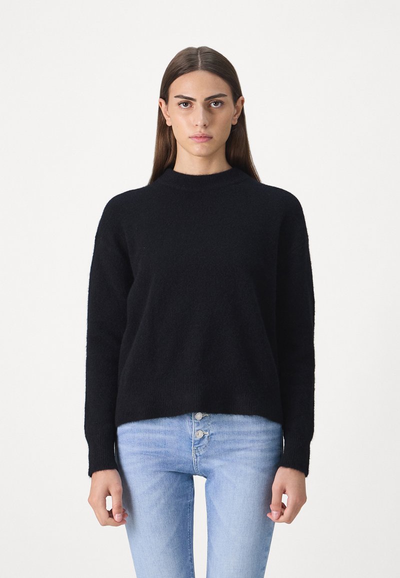 Schwarzer, flauschiger Pullover mit hohem Kragen und langen Ärmeln, kombiniert mit hellblauen Jeans mit Knopfverschluss.