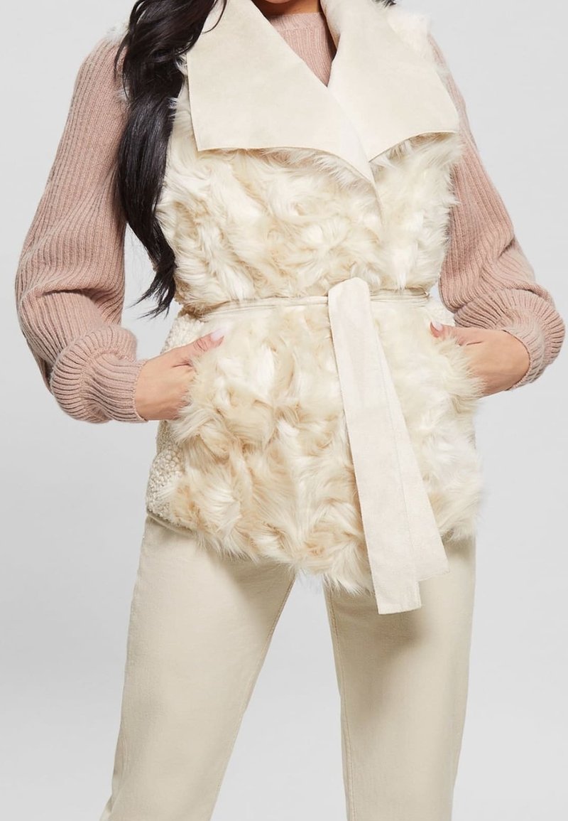 Donna che indossa un gilet in pelliccia sintetica beige con una cintura di tessuto, un maglione rosa a coste e pantaloni color crema, con le mani nelle tasche del gilet.