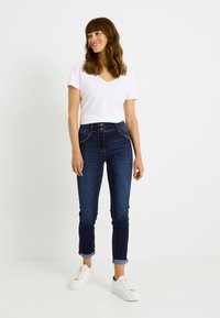 Camiseta blanca de manga corta con escote en V combinada con vaqueros de tiro alto azul oscuro con puños remangados y detalles de costura clara, complementada con zapatillas blancas.