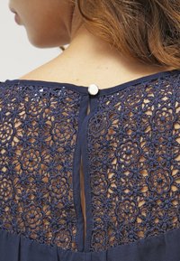 Abito blu navy con dettagli intricati in pizzo sul retro, chiusura con bottoni e orlo inferiore fluente.