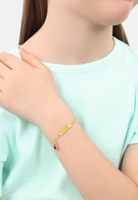 Bracciale d'oro con un ciondolo rettangolare piatto e un pendente di pietra rossa, caratterizzato da un design di catena attorcigliata. Indossato su un polso dalla pelle chiara.