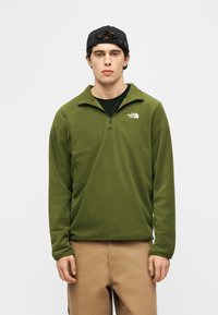 Νέος άνδρας που φοράει μαύρο καπέλο, πράσινο φλις με φερμουάρ και λογότυπο The North Face, μαύρο πουκάμισο από κάτω και μπεζ παντελόνι, στέκεται μπροστά από ένα απλό φόντο.