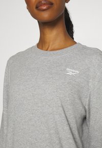 Sivi pamučni dukserica s okruglim izrezom, dugim rukavima i bijelim Reebok logotipom na gornjem lijevom dijelu. Glatka tekstura, ležeran dizajn.