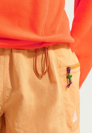 Haut en polaire orange rentré dans un pantalon orange clair avec une taille à cordon de serrage et une poche zippée ornée d'un petit tire-zip en forme de cactus vert.