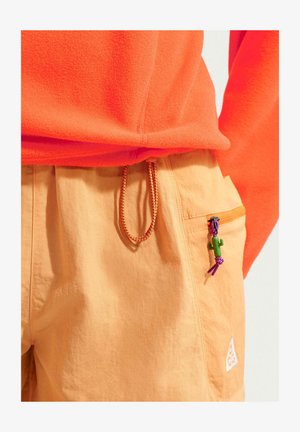 Narancssárga mikropolár felső világos narancssárga nadrágba tűrve, amelynek húzózsinóros derékrésze és cipzáras zsebe van, kis zöld kaktusz cipzárhúzóval.