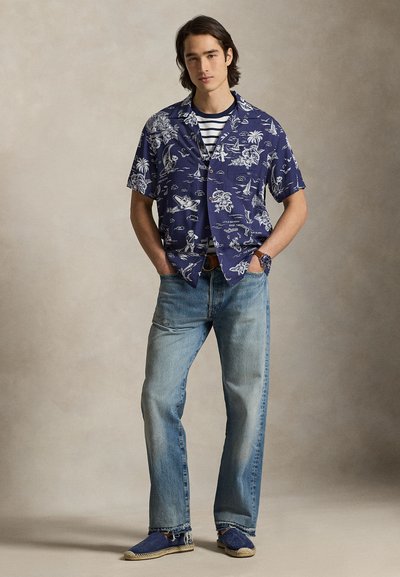 Młody mężczyzna w niebieskiej koszuli z krótkim rękawem w wzory, nałożonej na koszulkę w paski, jasnoniebieskich dżinsach i niebieskich espadrylach, stoi z rękami w kieszeniach.