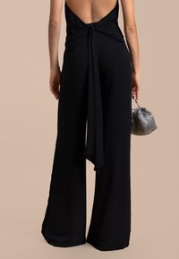 Uncombinaison noire avec un design sans dos, un détail noué à la taille, des jambes larges et une finition mate. Le mannequin tient un sac à main en argent.