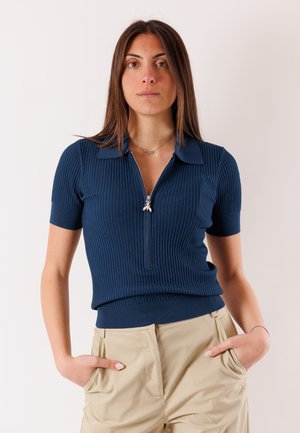Donna con una maglia a maniche corte a coste blu navy con cerniera frontale e pantaloni beige, in piedi con le mani nelle tasche contro uno sfondo semplice.