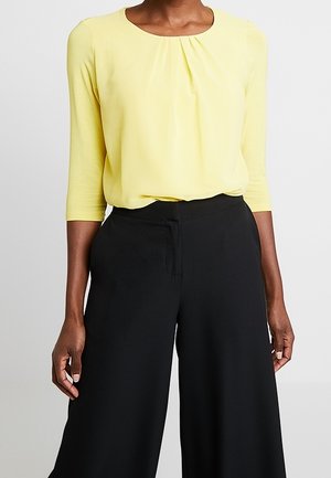 Gul bluse med lange ærmer og rynket halsudskæring, indsat i højtaljede, vidde, sorte bukser på en stående person.