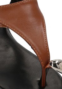 Sandalia de cuero marrón con una cinta superior curvada y un acento plateado con un extremo redondeado. Suela negra texturizada visible debajo de la cinta.