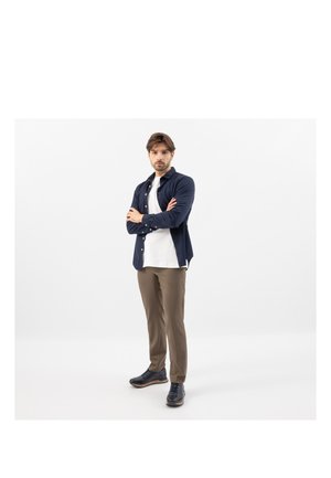 Man standing with arms crossed, wearing a navy shirt over a white t-shirt, brown pants, and black shoes, against a plain white background.