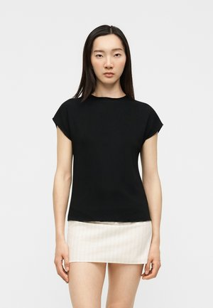 Femme debout portant un haut noir à manches courtes et une mini-jupe blanche avec des fines rayures verticales sur un fond uni.