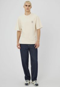 T-shirt en coton beige à manches courtes avec un détail de poche à l'avant à gauche, associé à un pantalon en denim bleu foncé avec des surpiqûres jaunes.
