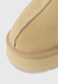 Tan suede footwear with a rounded toe, featuring visible stitching and textured rubber sole. Soft surface and smooth accents noted.