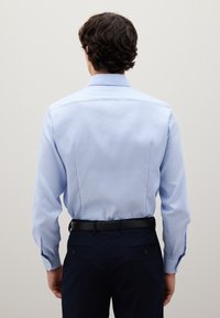 Uomo con capelli ricci scuri che indossa una camicia a maniche lunghe azzurro chiaro infilata in pantaloni blu scuro con una cintura nera, visto da dietro.