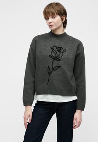 Jongere met kort bruin haar, gekleed in een donkergrijze trui met een zwart rozenontwerp, over een wit shirt en donkere spijkerbroek.