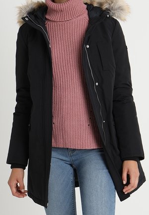 Parka noire avec une capuche en fausse fourrure, fermeture éclair, et poches avant, portée par-dessus un pull en maille rose et un jean bleu.
