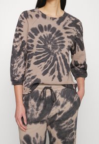 Person klädd i en beige och svart tie-dye sweatshirt och matchande sweatpants, med vänster hand i fickan mot vit bakgrund.