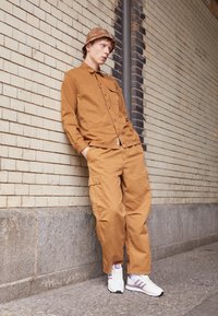 Brown cotton shirt and cargo pants, white sneakers with pink accents, tan patterned bucket hat. Person leans against a brick wall.