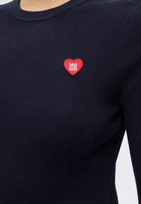 Gros plan sur un haut côtelé bleu marine avec un petit écusson en forme de cœur rouge sur la poitrine, brodé du mot "HUGO" en lettres blanches.