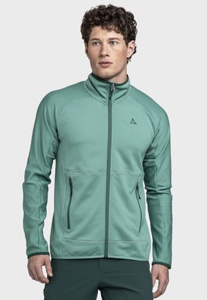 Mann mit lockigem Haar, der eine hellgrüne Jacke mit Reißverschluss und Reißverschlusstaschen trägt, vor einem schlichten hellgrauen Hintergrund steht.