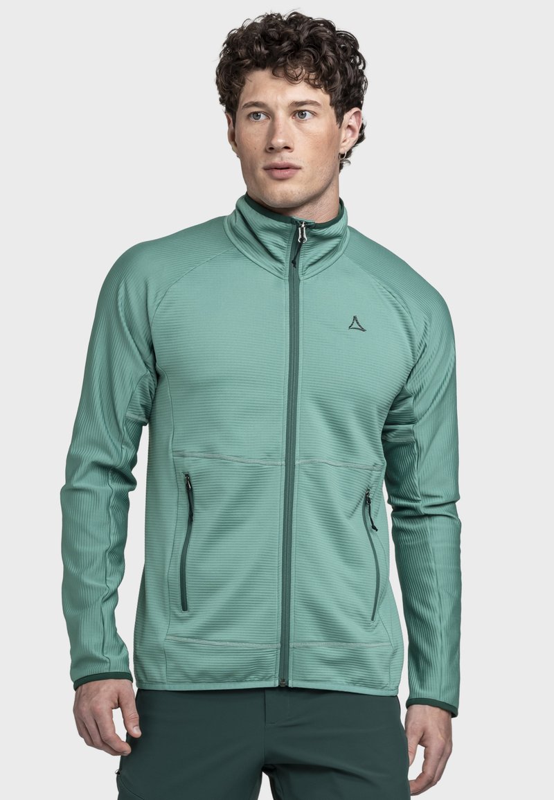 Mann mit lockigem Haar, der eine hellgrüne Jacke mit Reißverschluss und Reißverschlusstaschen trägt, vor einem schlichten hellgrauen Hintergrund steht.