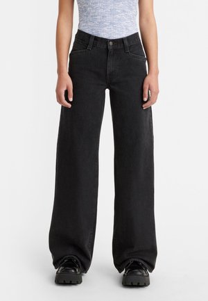 Personne portant un jean noir taille haute à jambes larges, rentré au-dessus de chaussures noires épaisses et associé à un haut bleu clair à motifs.