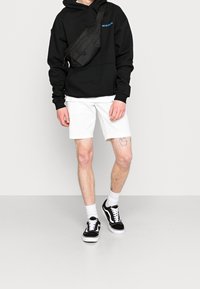 Felpa nera con testo blu, pantaloni corti bianchi e scarpe nere con suola bianca. Borsa a tracolla testurizzata con dettagli in zip.
