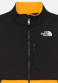 Svart fleecejacka med hög gul krage, framdragkedja och liten bröstficka med "The North Face"-logotyp i vitt.