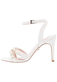 Sandale blanche à talons avec un matériau pailleté. Présente un design à lanières et des embellissements de perles et de pierres transparentes. Bride de cheville ajustable.