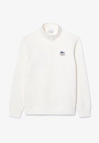 Bílý fleecový pulovr s vysokým límcem, půlzipem a tmavě modrým logem Lacoste na levé straně hrudi. Hladká textura a volný střih.