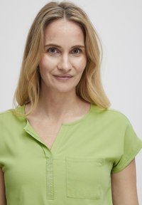 Blusa leggera verde chiaro a maniche corte e scollatura a V, con taschino sul petto e dettagli di cucitura a vista. Tessuto con consistenza liscia e vestibilità comoda.