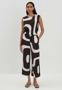 Zwart-witte mouwloze jumpsuit met abstract patroon, elastische taille, wijde pijpen en zijzakken. Gedragen met platte sandalen.