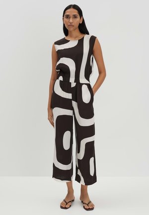 Schwarzer und weißer ärmelloser Jumpsuit mit abstraktem Muster, elastischem Bund, weit geschnittenen Beinen und seitlichen Taschen. Trägt flache Sandalen.